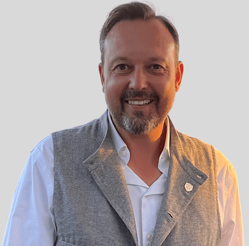 Man in a light gray vest and white shirt, smiling with a gray background.